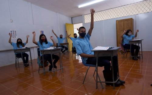 Varios estudiantes con mascarilla en un aula con las manos levantadas