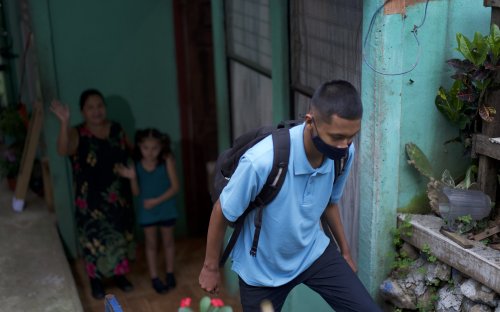 Estudiante con mascarilla saliendo de su casa, familia al fondo despidiéndose 
